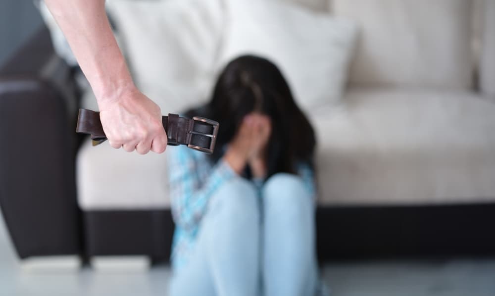 A man grips a belt in his fist while a woman cries in the background, illustrating the concept of domestic violence.