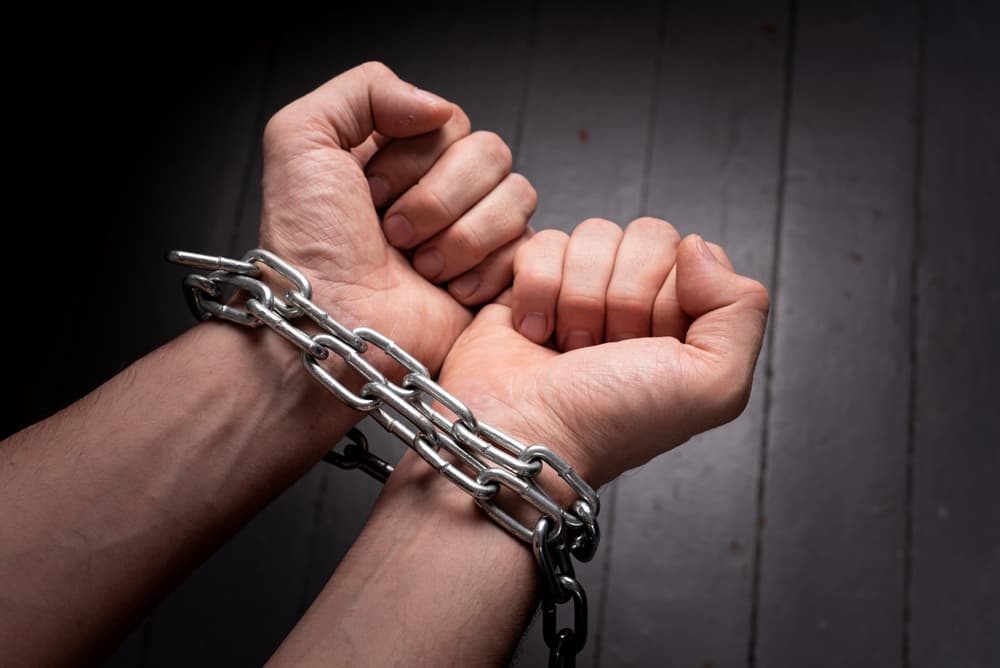 Male hands wrapped in a metal chain against a gray background, representing the concept of imprisonment.