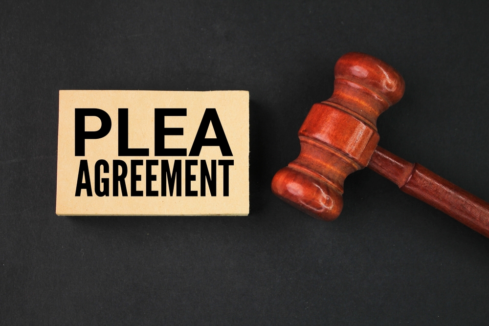 A wooden gavel next to a block labeled 'PLEA AGREEMENT' on a black background, symbolizing a legal plea deal or agreement in court proceedings.