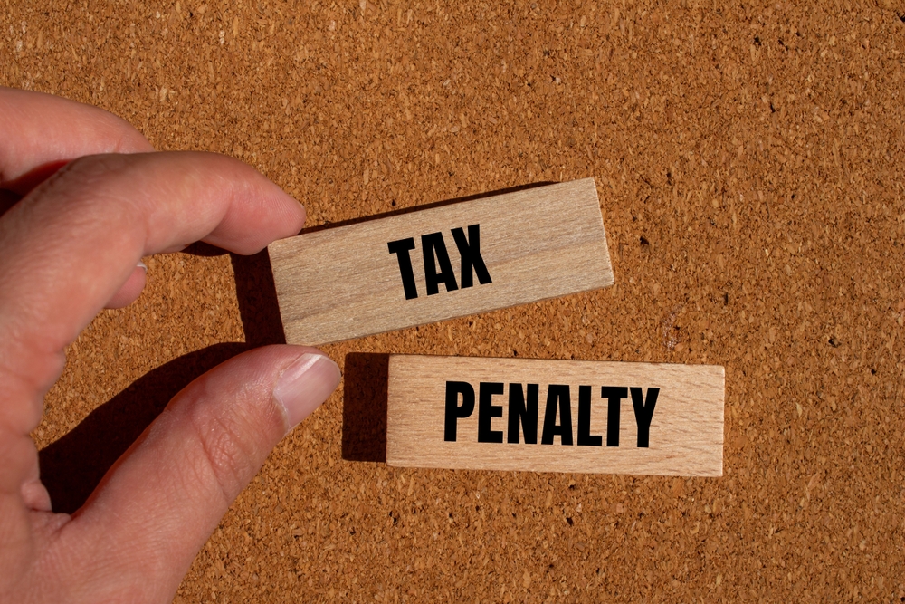 Hand holding wooden blocks labeled 'TAX' and 'PENALTY' on a corkboard background, symbolizing tax penalties.