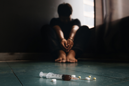 A person in handcuffs sitting on the floor in the background with a syringe and pills scattered on the floor in the foreground.