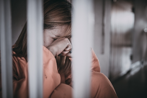 A distressed woman in an orange prison uniform sitting behind jail bars with her face in her hands.