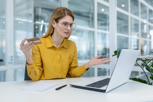 A concerned woman holding a credit card and looking at her laptop, representing frustration over potential online fraud.