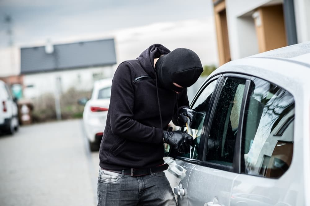 Masked thief attempting to pry open a car door with a screwdriver during a car theft attempt.